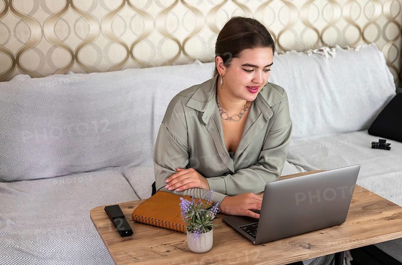 Young Caucasian woman with brown hair working on a laptop at home. She is smiling and sitting at a wooden table with a notebook and a plant