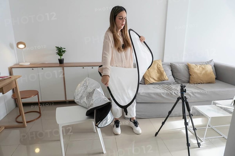 Woman standing and folding white photography diffuser reflector into black zippered carrying case, tripod and side table visible nearby