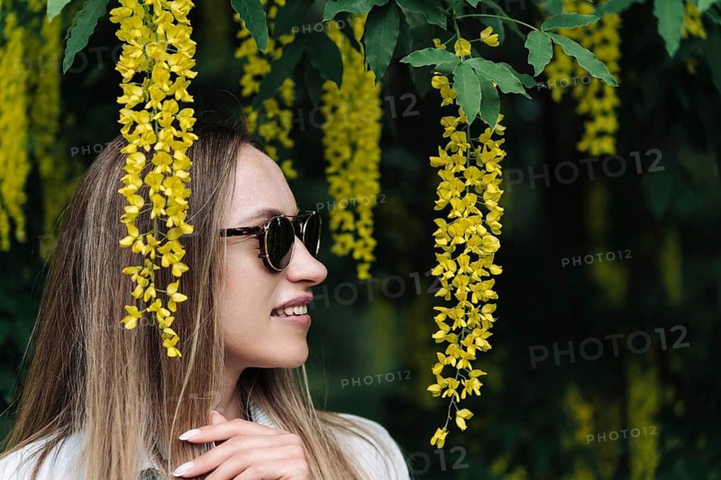 A woman walks among the blooming yellow flowers of Laburnum anagyroides, A woman wearing sunglasses smiles beside hanging yellow flowers in a lush, green outdoor setting