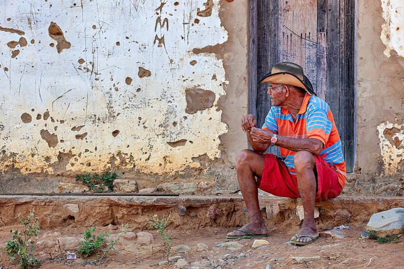 Elderly man sits thoughtfully in front of a faded wall on a village path, a man sits on a wall in the Caatinga of Bahia in Brazil