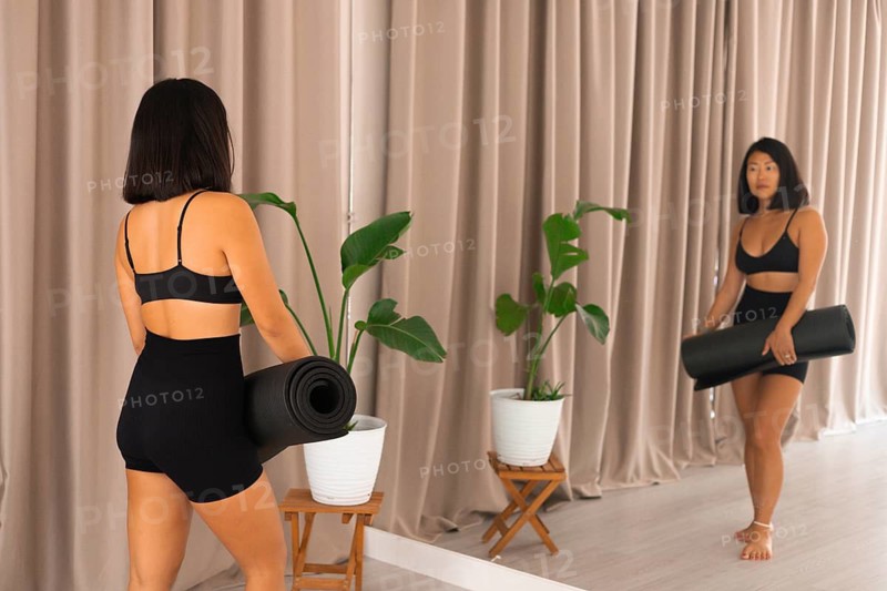 A woman holding a yoga mat looking at her reflection next to potted plants