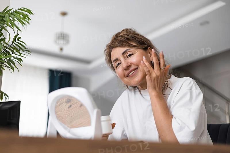A woman smiles as she applies a skincare product to her face in a well-lit indoor space. The natural light enhances her joyful expression while she interacts with a mirror. Morning routine