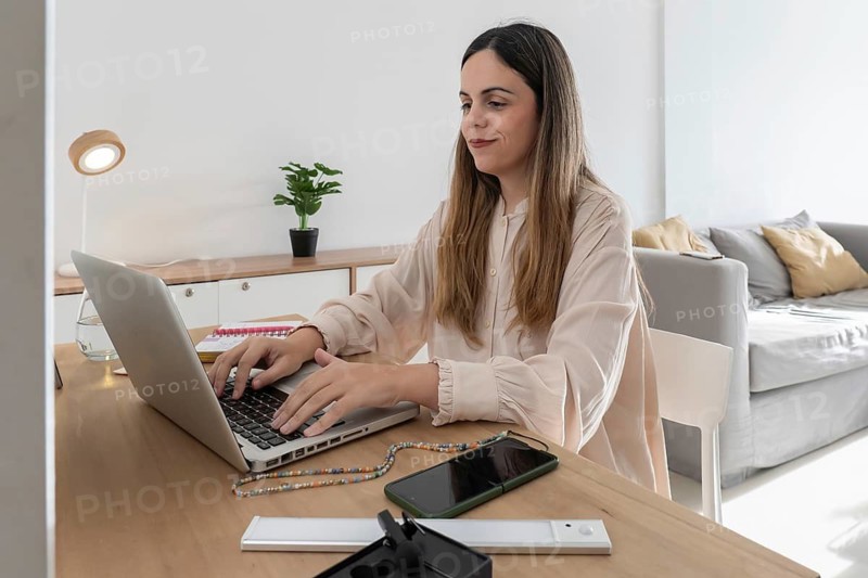 A content creator working at a wooden desk, typing on a laptop with a smartphone and headphones on the desk, in a bright, minimalist flat
