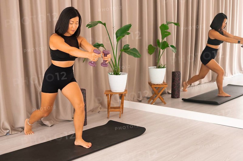 A woman lunges forward on an exercise mat with dumbbells, concentrating in an indoor setting