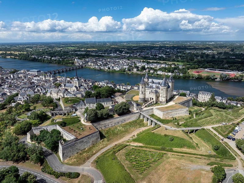 Aerial view of the historic Château de Saumur with its distinctive round corner towers and bright facades, surrounded by well-kept green spaces under a slightly cloudy summer sky, with the town of Saumur on the Loire River, Maine-et-Loire, Pays de la Loire, France