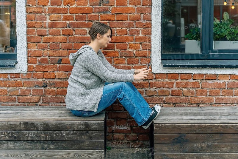 Woman 40s in knit cardigan using smartphone on wooden bench at brick building, A woman in casual clothing sits on a wooden bench against a brick wall, using her smartphone