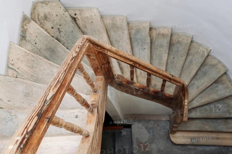 The steps of a wooden spiral staircase in an old building. An old wooden staircase in an old building with white walls, A spiral staircase with a wooden railing and concrete steps, creating a rustic and minimalist atmosphere