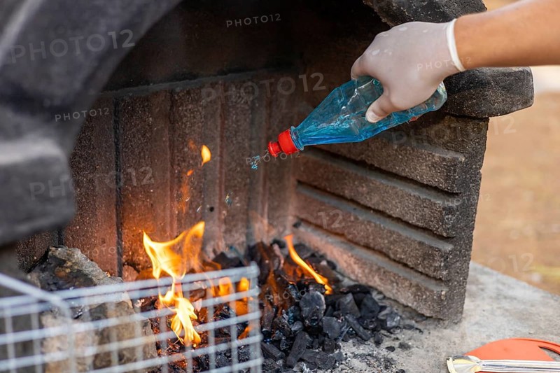 A gloved hand pours blue liquid onto burning charcoal in a stone barbecue grill. Flames rise from the coals, indicating preparation for cooking