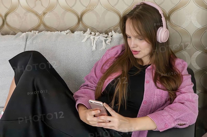 Young person sits on couch wearing headphones while using smartphone. The background shows a decorative wall and soft pillows. Sunlight fills the room in the afternoon