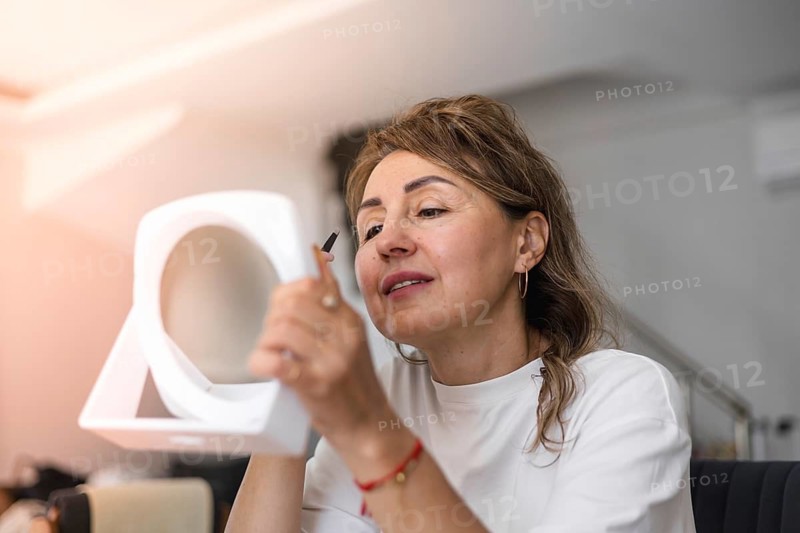 A woman sits in a stylish room, using a compact mirror, tweezers and doing eyebrow hair removal. Natural light illuminates her focused expression as she enhances her makeup routine with precision