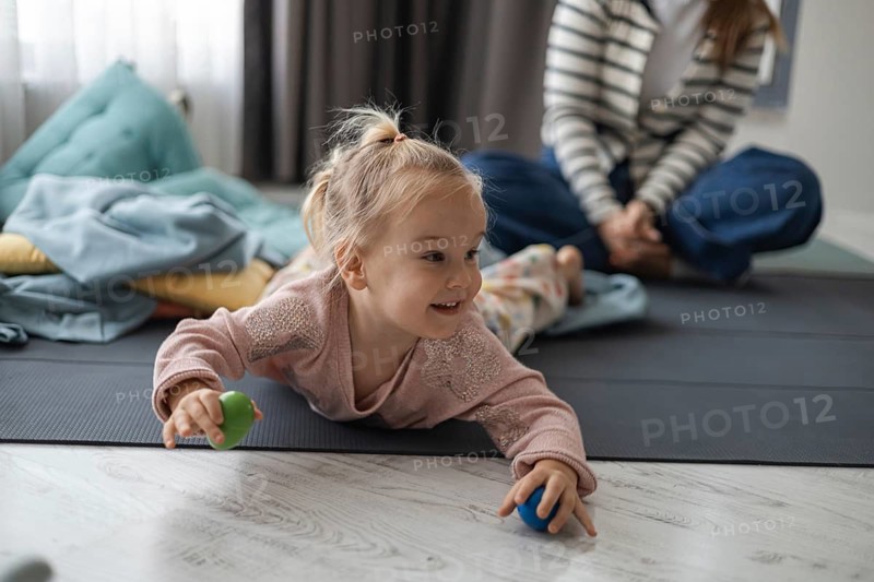 A young Caucasian girl with blonde hair plays on the floor with colorful balls. A woman in a striped shirt sits nearby, creating a cozy home atmosphere