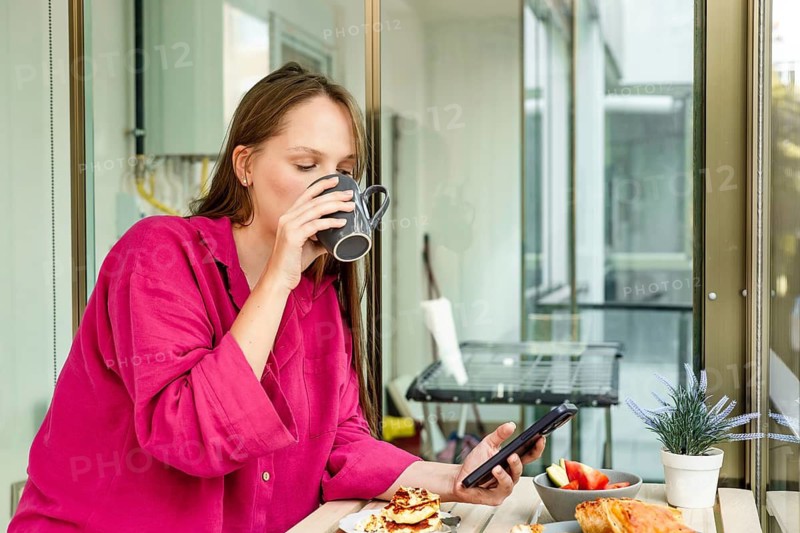 A young Caucasian woman with long brown hair drinks coffee while using a tablet. She wears a pink shirt and is seated at a table with breakfast items