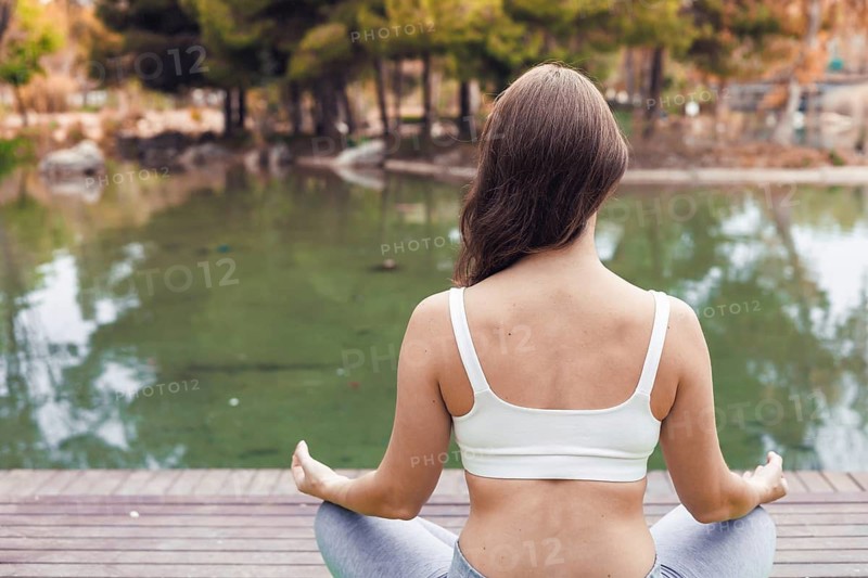 A young Caucasian woman with long brown hair sits cross-legged by a serene lake, practicing meditation in a peaceful outdoor setting surrounded by trees