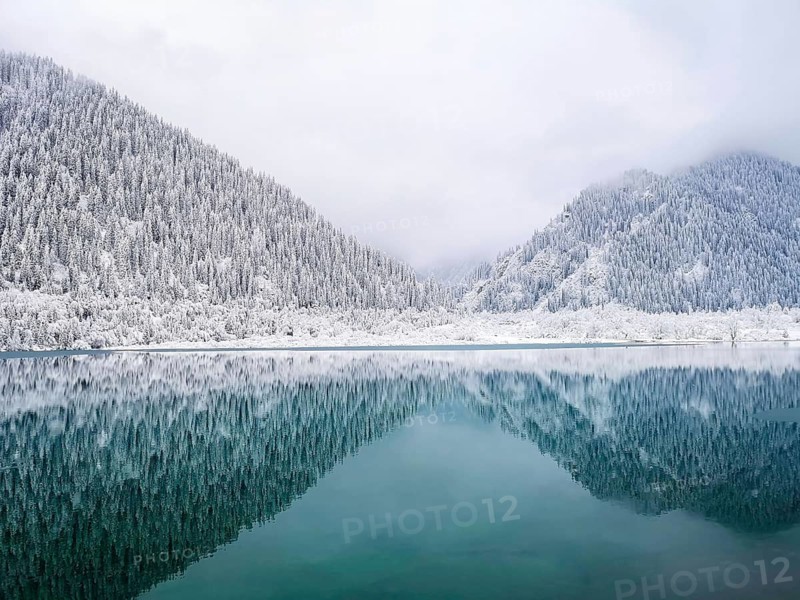 Mountains covered with trees and snow are symmetrically reflected in the turquoise water of the lake. Winter landscape, Snow-covered mountains reflected in a tranquil lake under a cloudy sky, Lake Issyk, Kazakhstan