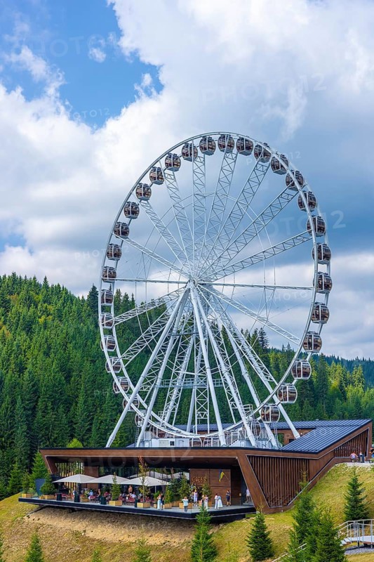 A large Ferris wheel set against a forested hillside under a partly cloudy sky