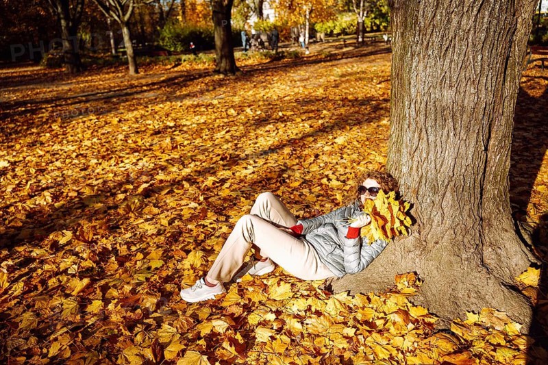 Woman relaxing with bouquet of maple leaves in sunny autumn park. A person reclines against a tree in a park covered with dense autumn leaves. Bright yellow foliage is held close to the face, creating a playful seasonal gesture. Light pants, a gray jacket, red gloves, and sunglasses add contrast to the warm fall colors. The scene conveys relaxation, cozy outdoor mood, autumn texture, and mindful enjoyment of nature during the season
