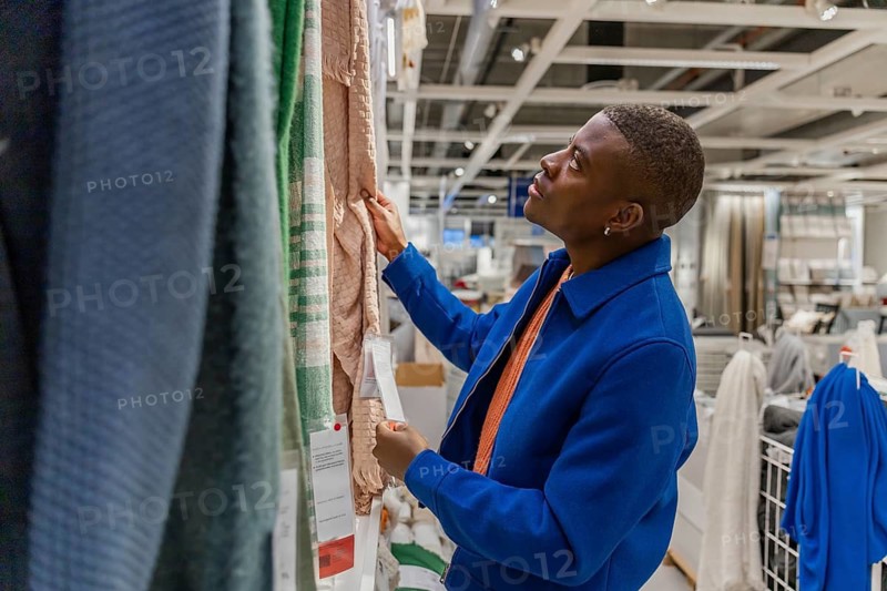 A young Black man examines fabric samples in a home goods store. He wears a blue coat and looks thoughtfully at the orange material