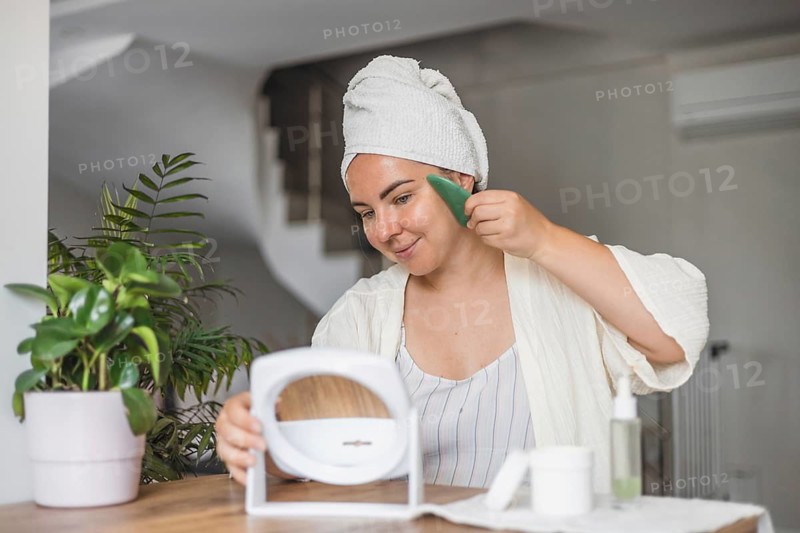 A young Caucasian woman with brown hair in a towel smiles while applying gua sha on her face. A potted plant and beauty products are on the table