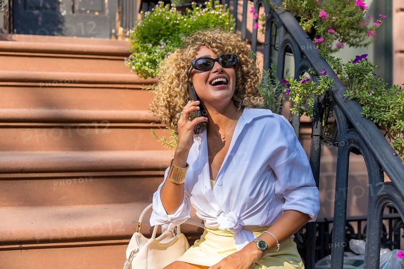 Young woman with curly blonde hair smiling and laughing while talking on her phone, sitting on brownstone steps in manhattan, embodying urban life and communication