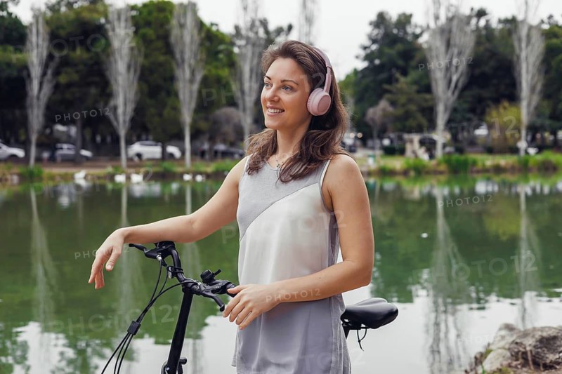 A young Caucasian woman with brown hair stands beside a bicycle near a serene lake. She wears pink headphones and smiles, enjoying the outdoors