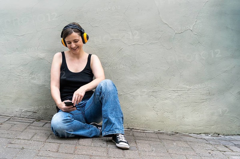 Smiling blonde woman 40s with phone and yellow headphones sitting on urban pavement, Woman sitting against a wall wearing yellow headphones, using a smartphone with a cheerful expression