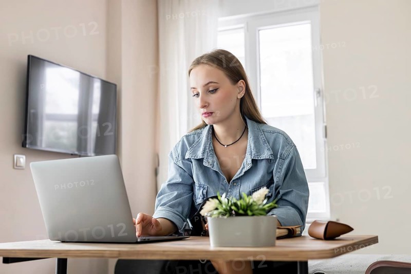 Young Caucasian woman with long brown hair working on a laptop at a wooden table. A potted plant is present. Bright indoor setting with a TV in the background