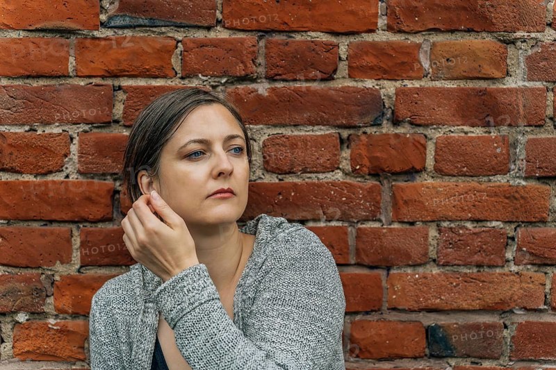 Thoughtful woman 40s with light blonde hair against red brick wall portrait, A woman adjusts her earring while sitting against a brick wall, appearing thoughtful and casual