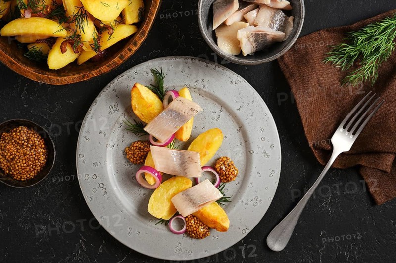 Plate with potato wedges alongside slices of herring. Small bowls hold mustard and fresh herbs. This scene is set on a dark surface creating a simple meal setup
