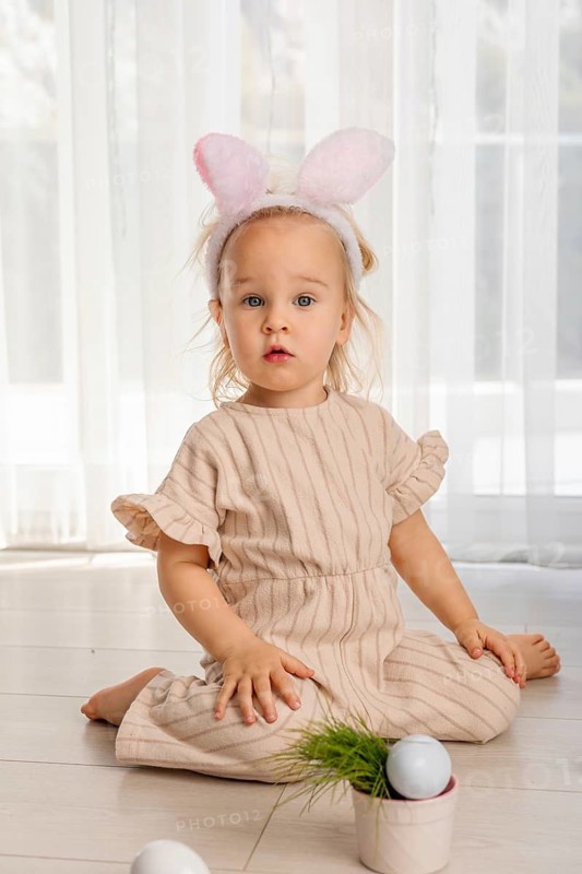 A toddler girl sits on the floor in a bright room. She wears bunny ears and smiles while surrounded by Easter eggs and a small plant. She enjoys the Easter holiday fun and celebration