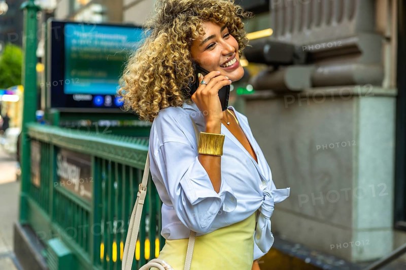 Happy young woman with curly hair talking on a cell phone, smiling in front of a new york city subway station entrance, embodying urban lifestyle