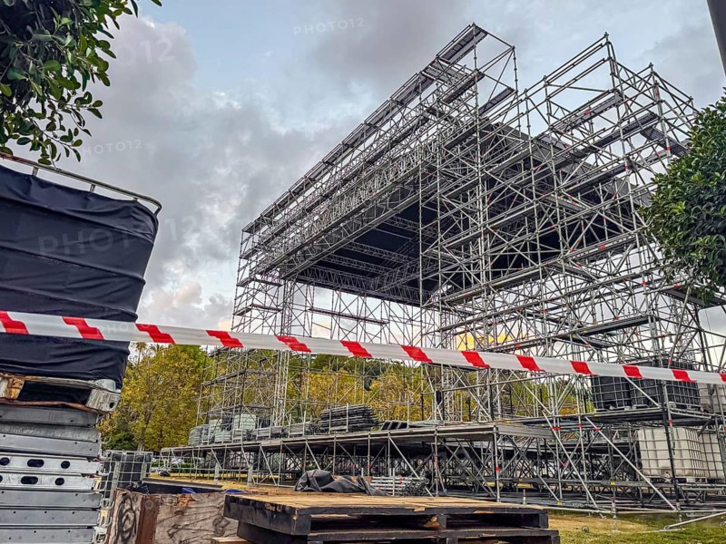 Construction site with scaffolding and safety barriers. Metal framework for a stage under assembly. Trees and cloudy sky in the background