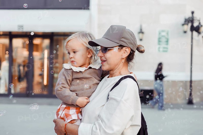 A woman holds her young daughter close as they watch bubbles float in the air. The city square is bustling with a warm autumn atmosphere, creating a joyful scene of bonding