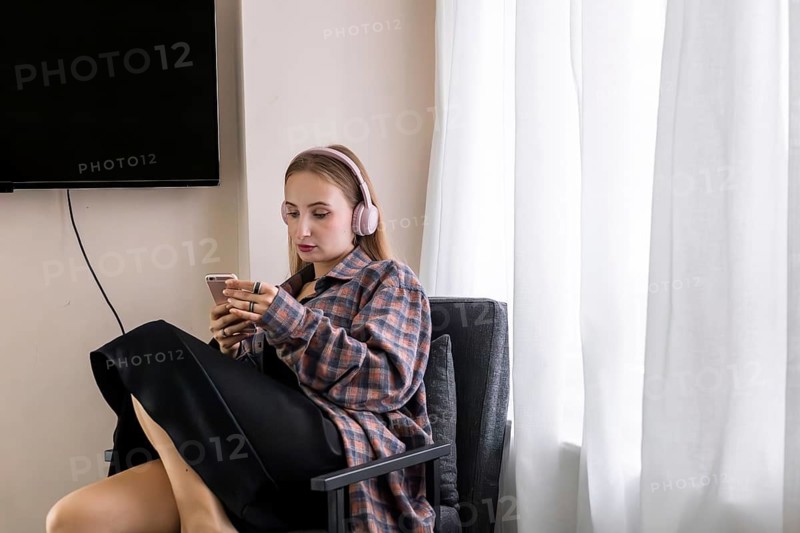 Young Caucasian woman with long brown hair, wearing headphones, sitting in a chair and using a smartphone in a bright room with a TV and curtains