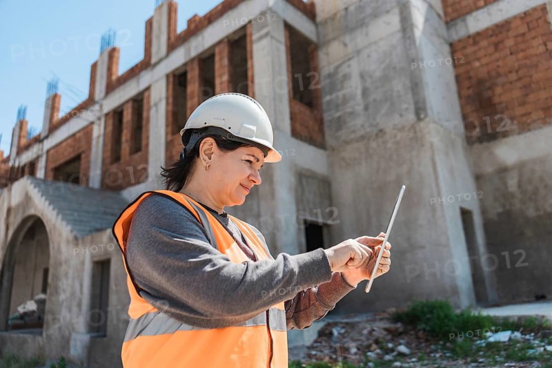 A young Hispanic woman in a hard hat and orange vest uses a tablet at a construction site. The background features a partially built structure