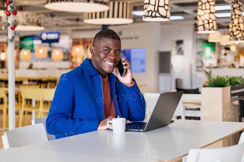Smiling young African man in a blue coat talking on a smartphone while sitting at a table with a laptop and a cup in a modern cafe