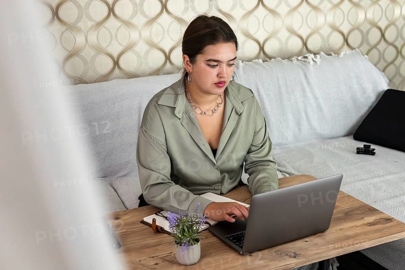 A young Hispanic woman with long brown hair works on a laptop at a wooden table in a cozy living room. A small plant is on the table
