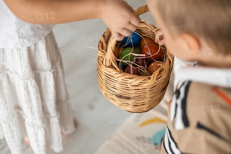 A young boy with short hair reaches into a woven basket held by a girl in a white dress. The scene captures a warm, family atmosphere at home. Easter eggs