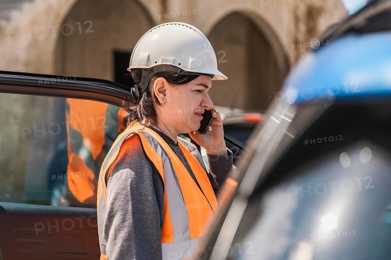 Worker woman in a hard hat and safety vest talks on her phone near a blue car. The scene is set outdoors with a clear sky