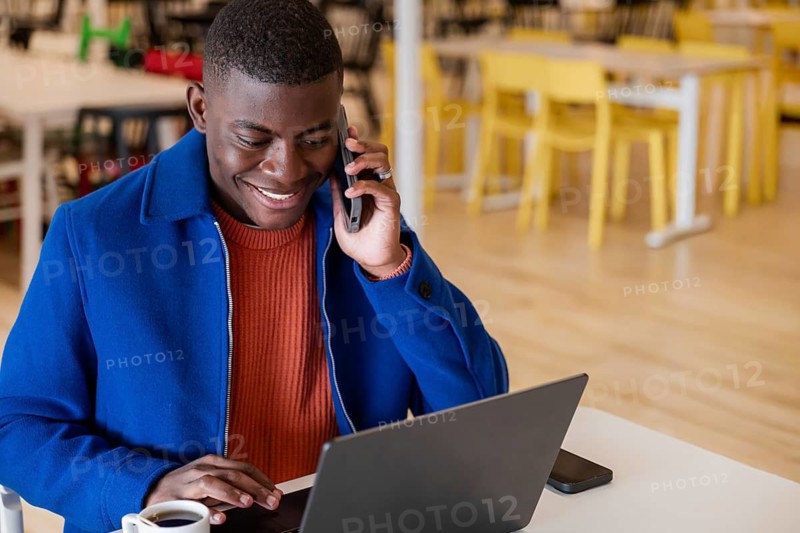 A young African man with short hair is sitting at a table in a cafe. He is smiling while talking on a smartphone and using a laptop. A cup is on the table