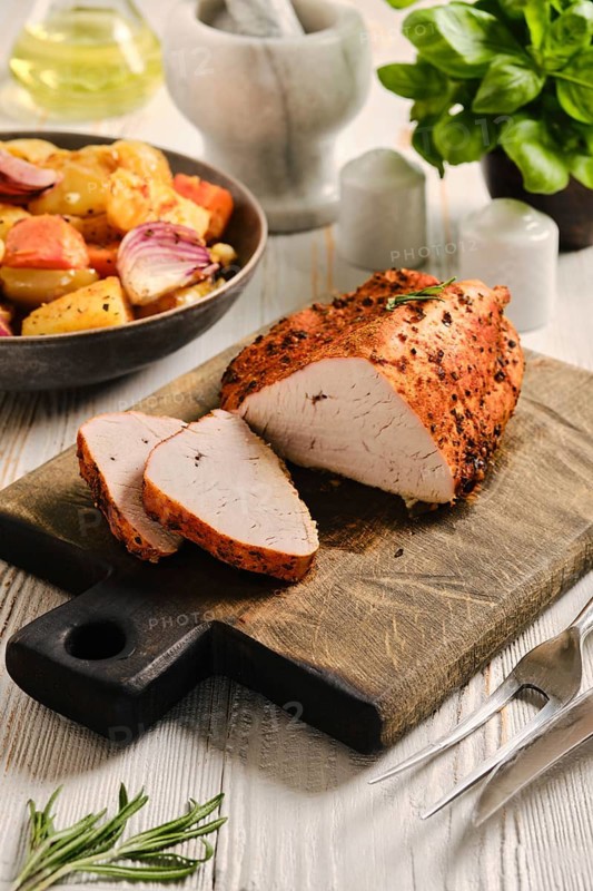 Roasted turkey breast on wooden cutting board. Slices of turkey meat are arranged next to whole piece. Roasted vegetables are in bowl nearby ready for serving