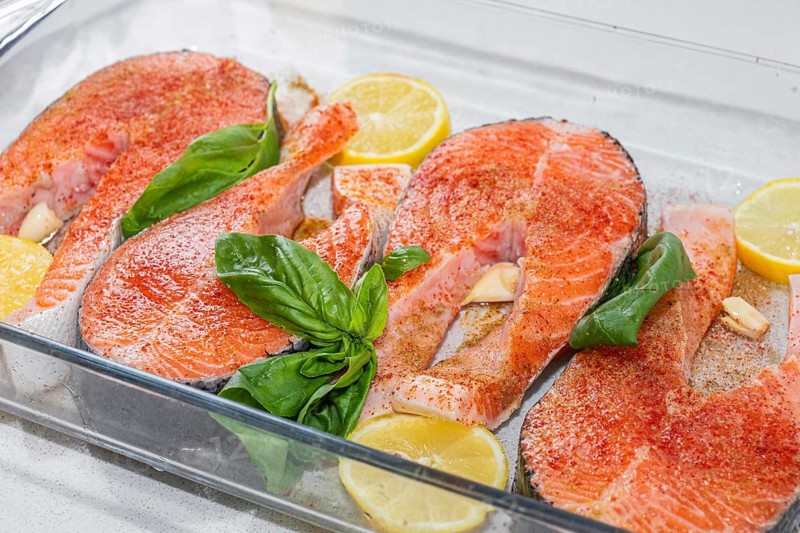 Fresh salmon fillets arranged in a glass dish with lemon slices and basil leaves. The fish is raw and ready for cooking. Healthy food and diet concept