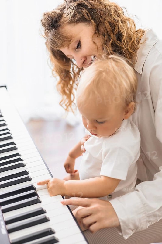 A woman gently teaches a baby at the piano, pointing at the keys with care
