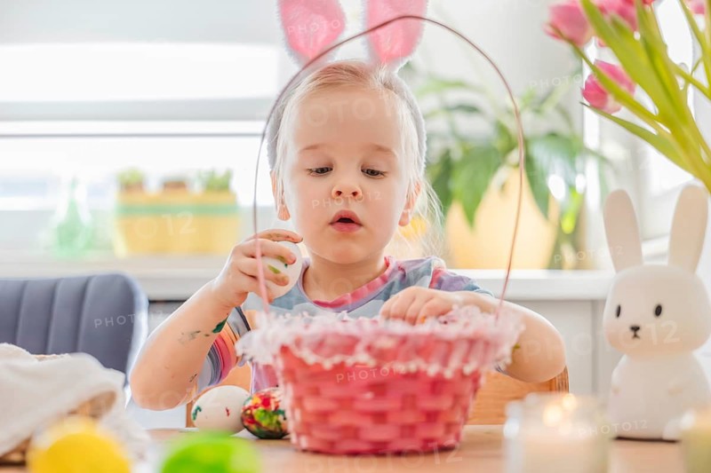 A young child wears bunny ears and dips an Easter egg into a basket at a table. Flowers and candles are around, creating a fun atmosphere for holiday crafts