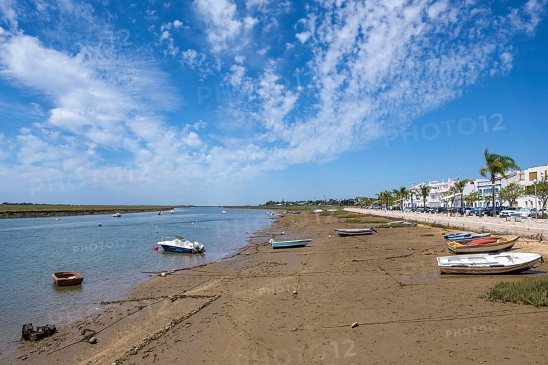Boats lie on the sandy shore of a lagoon with blue skies and scattered clouds, flanked by white buildings and palm trees, Santa Luzia, Tavira, Faro, Portugal