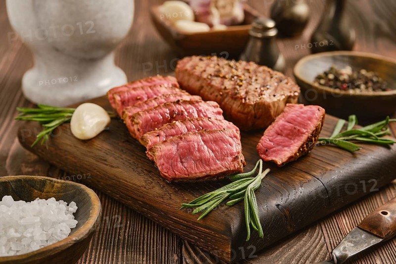 Beef steak is sliced and displayed on wooden board. Whole garlic cloves and bowls of spices are nearby. The setting is a kitchen or dining area with wooden surfaces