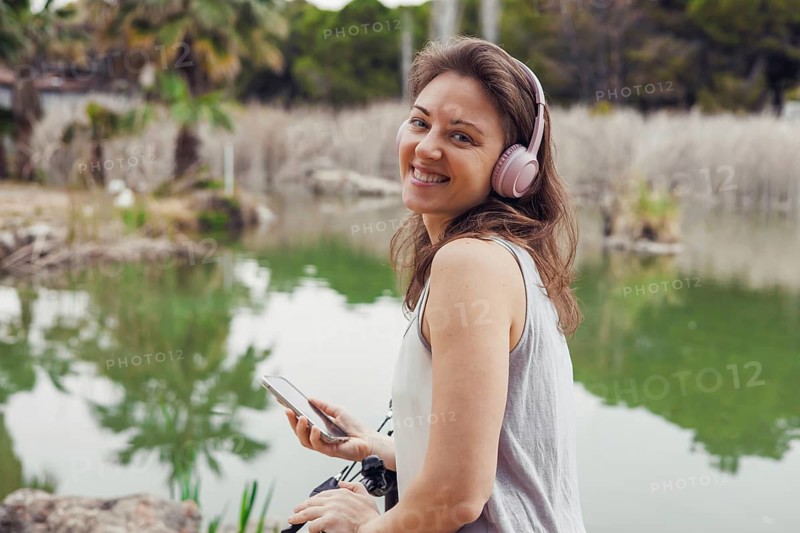 A young Hispanic woman with long brown hair smiles while wearing headphones. She holds a smartphone near a tranquil pond surrounded by greenery