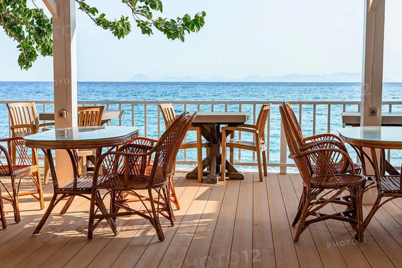 Rattan chairs and tables set on a seaside terrace overlooking clear blue water on a bright and sunny day