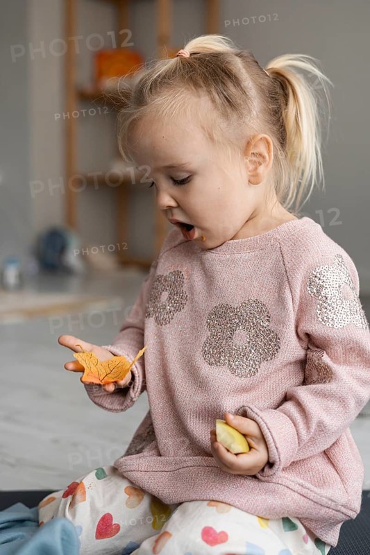 A young Caucasian girl with blonde hair in pigtails sits on the floor, holding orange leaves. She appears curious and engaged in a sensory activity at home
