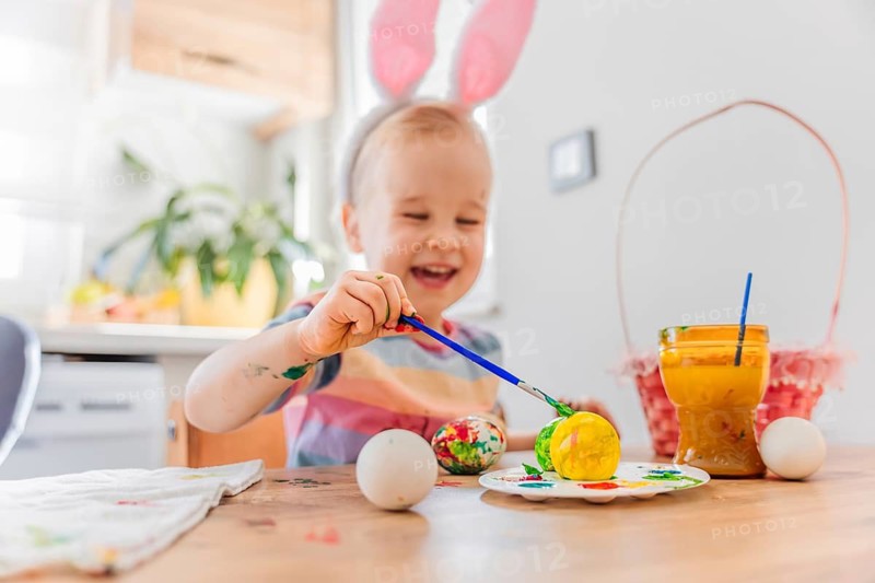 A child wearing bunny ears sits at a table decorated for Easter. Colorful eggs are placed on the table. The room has bright light and festive items. The child looks cheerful and excited