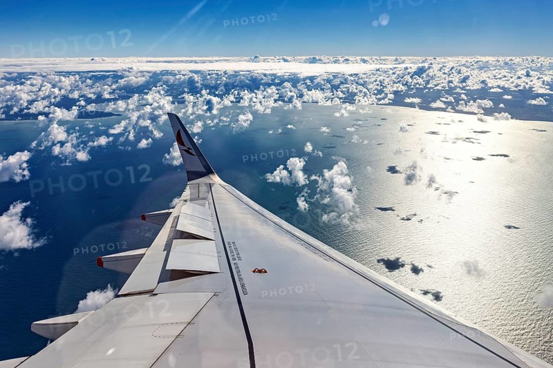 View from an airplane window of the ocean with clouds and sunlight reflecting water, aircraft wings above the clouds
